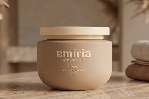 A beige cosmetic jar labeled 'emiria' sits on a stone countertop in a softly styled bathroom setting with folded towels and dried flowers in the background.