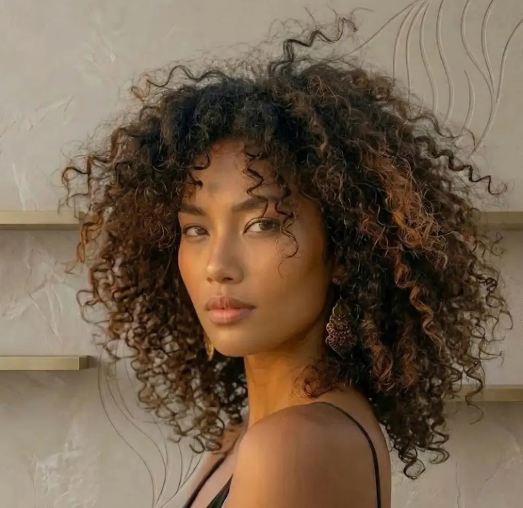 A close-up portrait of a woman with voluminous, tightly curled dark brown hair, wearing a black spaghetti-strap top and a dangling earring, set against a textured beige wall with light wood shelves.